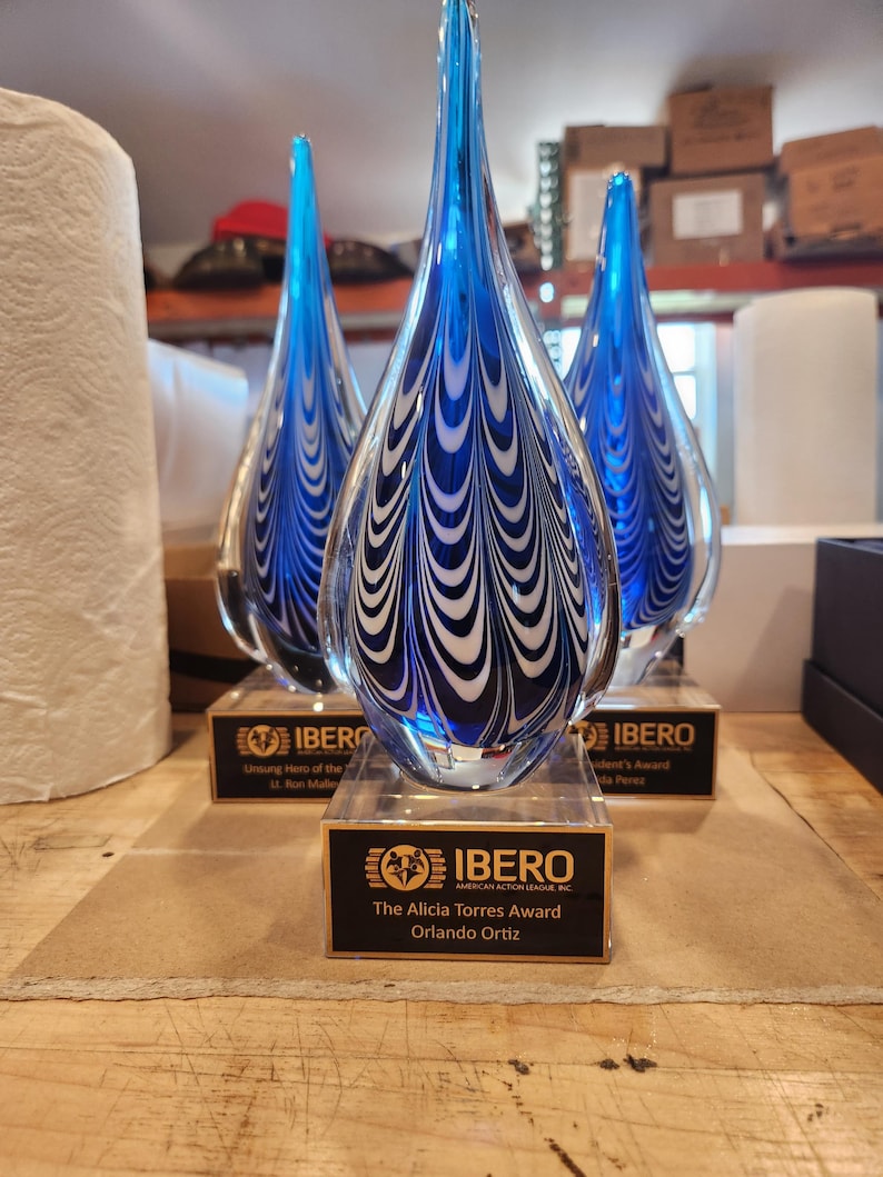 May include: Three teardrop-shaped glass awards with blue and white swirling patterns. Each award sits on a clear rectangular base with a black plaque. The front plaque reads: IBERO AMERICAN ACTION LEAGUE, INC. The Alicia Torres Award, Orlando Ortiz.