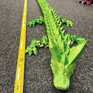 May include: A bright green, articulated dragon toy, approximately 54 inches long, lies on a gray surface. The dragon is made of geometric shapes, with a spiky spine and detailed head. A yellow measuring tape is next to the dragon.
