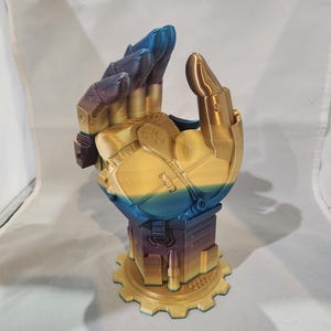 May include: A 3D printed gold and blue hand sculpture with a gear base. The hand is posed in a fist with the thumb and index finger extended. The sculpture is made of a smooth, shiny material.