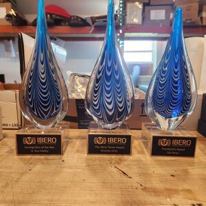 Three hand-blown blue and black art glass teardrop awards with engraved nameplates on wood bases, custom corporate recognition trophies displayed on a workshop table.