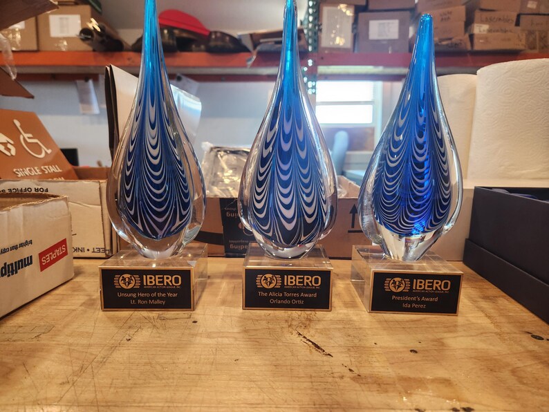 Three hand-blown blue and black art glass teardrop awards with engraved nameplates on wood bases, custom corporate recognition trophies displayed on a workshop table.