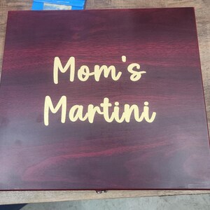 May include: A rectangular wooden box with a dark burgundy finish. The words "Mom's Martini" are written in a gold-colored script across the top of the box. The box appears to be closed.