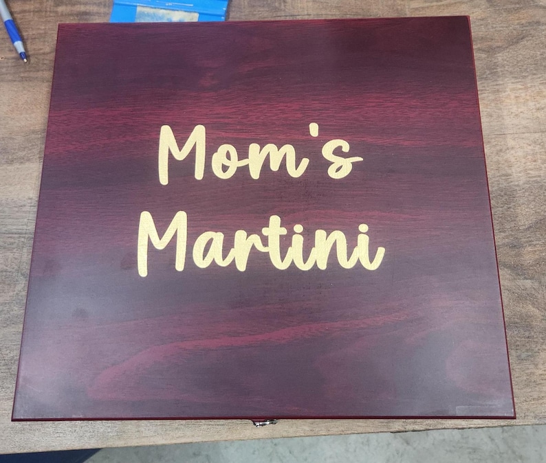 May include: A rectangular wooden box with a dark burgundy finish. The words "Mom's Martini" are written in a gold-colored script across the top of the box. The box appears to be closed.