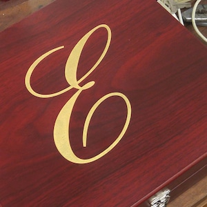 Close-up of a personalized rosewood finish gift box featuring an engraved and gold color-filled script initial 'E'. The elegant monogram design is centered on the box lid, making it ideal for a custom cocktail gift set, barware set, or special occasion present. This engraved wood box offers a luxurious presentation and is perfect for weddings, groomsmen, Father's Day, anniversaries, or corporate gifting.