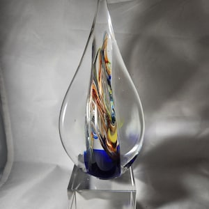 May include: A teardrop-shaped clear glass sculpture featuring an internal swirl of blue, yellow, orange, and white colors. The sculpture is mounted on a solid, rectangular clear glass base, creating a smooth, reflective, and transparent decorative object.