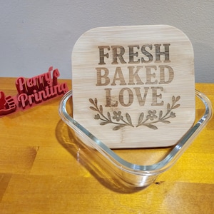 May include: A clear glass baking dish with a wooden lid. The lid is engraved with the words "FRESH BAKED LOVE" and a floral design. A red 3D printed logo is in the background.