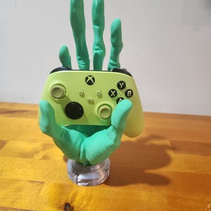 May include: A green 3D printed hand holding a lime green Xbox controller. The hand is holding the controller in a relaxed, open-handed position. The controller is resting on the palm of the hand. The hand is attached to a gray base.