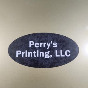 May include: An oval-shaped sign with the text "Perry's Printing, LLC" in white lettering against a dark, textured background. The sign is set against a gold-colored surface.
