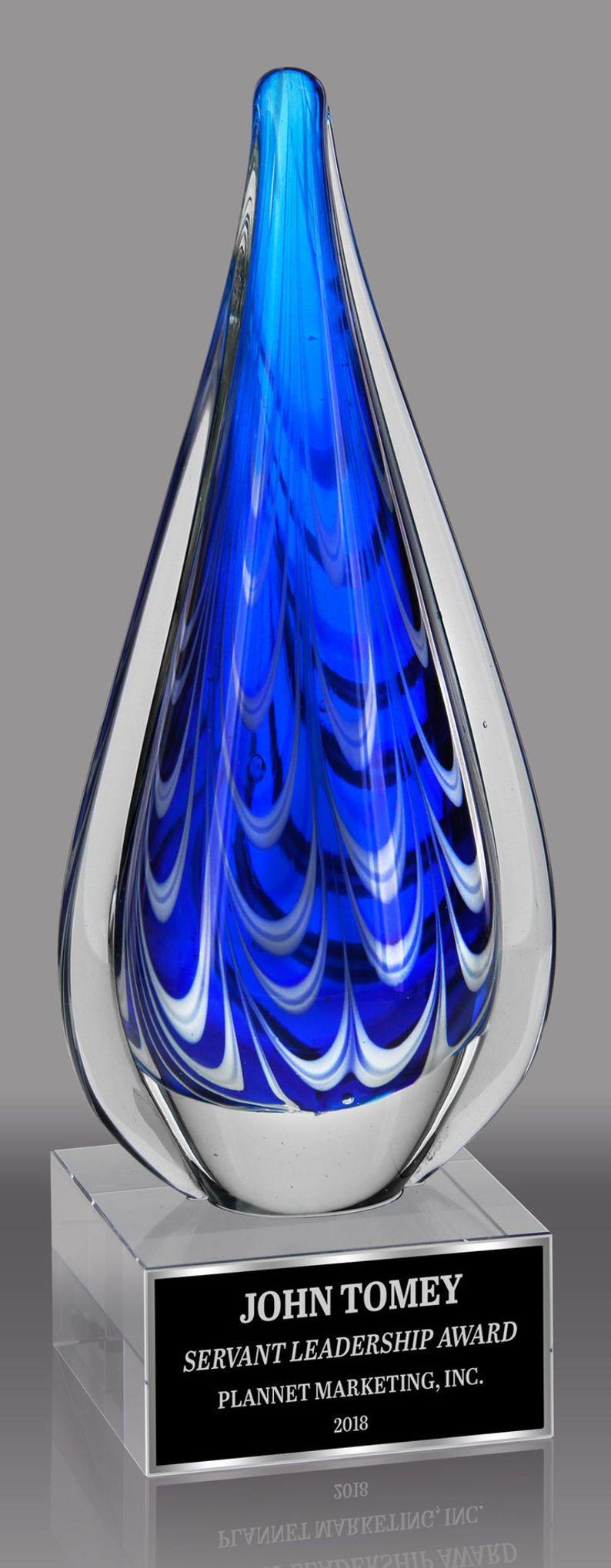 A blue glass award with white swirls. The award is on a clear acrylic base with a black plaque that reads 'JOHN TOMEY SERVANT LEADERSHIP AWARD PLANNET MARKETING, INC. 2018'.
