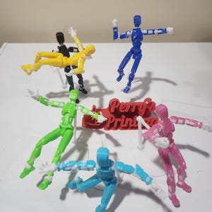May include: A collection of articulated action figures in various colors, including yellow, blue, green, pink, and black. Each figure is posed in a dynamic stance, with white hands and feet. The figures are on white stands, with a red sign that reads "Perry's Printing".