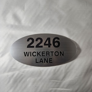 May include: Silver oval-shaped house number sign with black text. The number 2246 and the street name "Wickerton Lane" are printed on the sign.