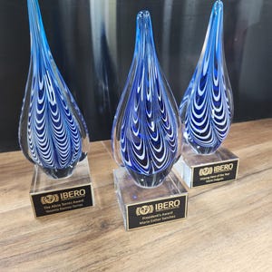 Set of three artisan glass teardrop awards featuring stunning blue and white swirl patterns, mounted on clear rectangular bases with custom engraved nameplates. Ideal for corporate recognition, appreciation ceremonies, or employee achievement awards. Elegant design makes each piece a unique and memorable gift or display item for honoring excellence and dedication. Perfect for office décor or award showcases.