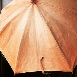May include: A vintage-style umbrella with a light brown fabric canopy. The umbrella is open, showcasing its radiating panels and a decorative top. A matching strap hangs from the handle. The umbrella's design suggests a classic, possibly antique, aesthetic.