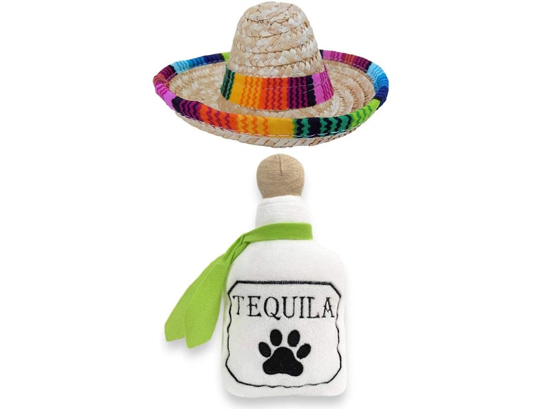 Op de afbeelding: Een witte pluche tequilafles met een groene sjaal en een zwarte pootprint. De fles heeft het woord "Tequila" geborduurd. Een strooien sombrero met een kleurrijke band is ook inbegrepen.