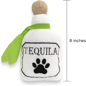 Op de afbeelding: Wit pluche hondenspeelgoed in de vorm van een tequilafles met een groen lint en een zwarte pootprint. De fles is 20 cm hoog en heeft het woord "TEQUILA" op het etiket.