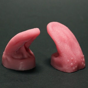 Puede incluir: Dos objetos rosas con forma de lengua y superficies texturizadas. Los objetos son de color rosa claro y tienen un aspecto realista, con pequeños bultos y pliegues. Se encuentran sobre un fondo oscuro.