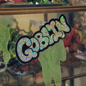 May include: A sticker with the word "CABAL" in a bubble letter style, outlined in black, with a holographic, glittery fill. The sticker is displayed in front of a glass shelf with other figurines.