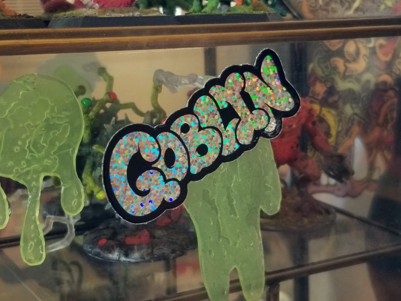 May include: A sticker with the word "CABAL" in a bubble letter style, outlined in black, with a holographic, glittery fill. The sticker is displayed in front of a glass shelf with other figurines.