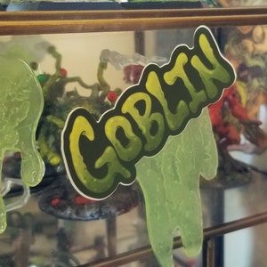 May include: A sticker with the word "GOBLIN" in a green and yellow gradient font. The sticker is placed in front of a display case with green slime and other fantasy-themed objects.