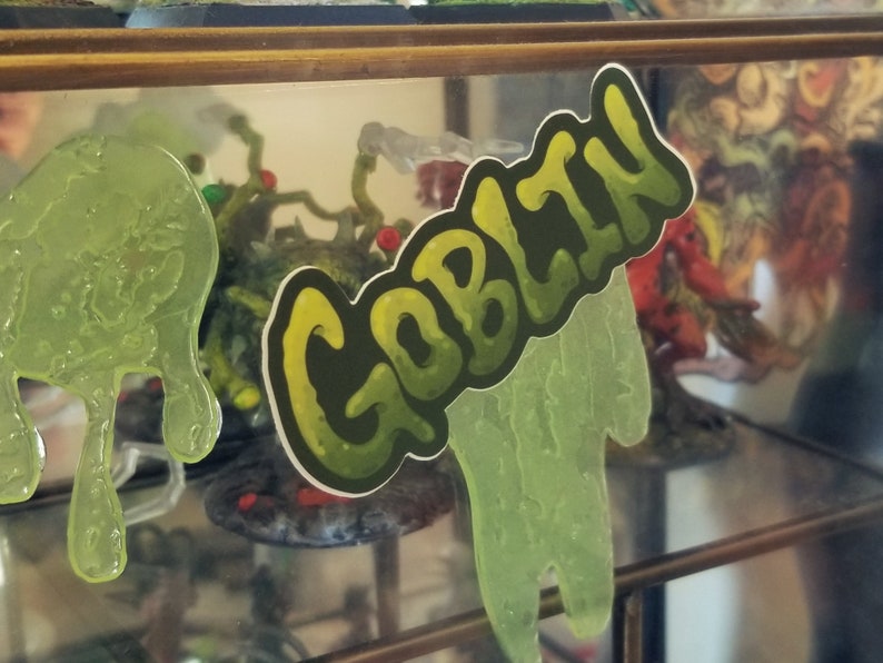 May include: A sticker with the word "GOBLIN" in a green and yellow gradient font. The sticker is placed in front of a display case with green slime and other fantasy-themed objects.