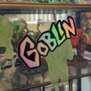 May include: A sticker with the word "GOBLIN" in colorful, outlined letters. The sticker has a holographic effect and is placed over a green, drippy, slime-like shape. The background includes miniature figurines.