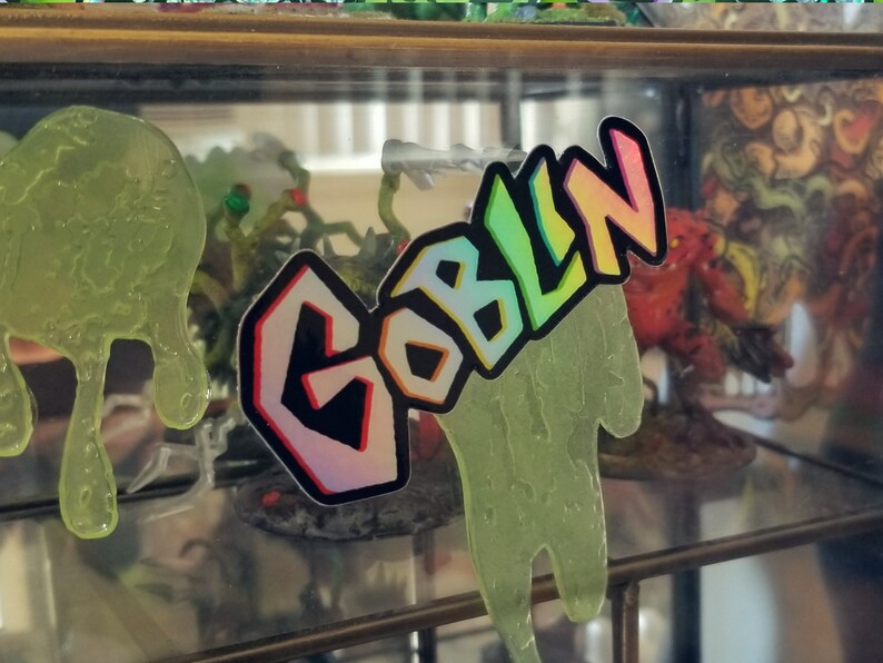 May include: A sticker with the word "GOBLIN" in colorful, outlined letters. The sticker has a holographic effect and is placed over a green, drippy, slime-like shape. The background includes miniature figurines.