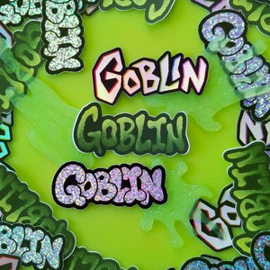 May include: A collection of stickers with the word "Goblin" in various styles. The stickers feature a mix of colors, including green, white, and iridescent accents. Some stickers have a glittery finish, and the background is a bright green.