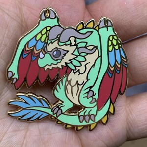 May include: A green and gold enamel pin of a cartoon dragon with red, blue, and yellow feathers. The dragon has a light brown body and a light gray horn.