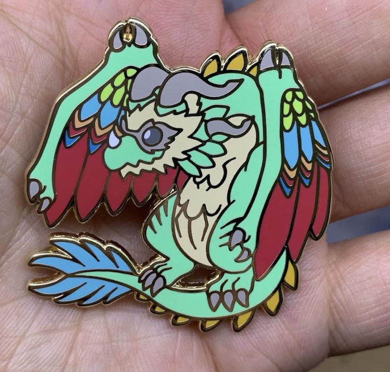 May include: A green and gold enamel pin of a cartoon dragon with red, blue, and yellow feathers. The dragon has a light brown body and a light gray horn.