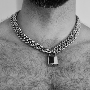 Peut inclure: Un collier en chaîne argentée avec un pendentif en forme de cadenas. Le collier est porté par une personne avec des poils sur la poitrine.