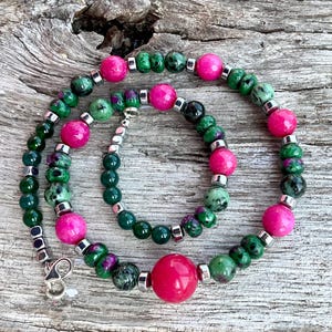 Può includere: Una collana con una combinazione di perline rosa e verdi. La collana presenta un mix di perline rotonde e più piccole, con distanziatori argentati. La chiusura è argentata. Le perline sono disposte su una superficie di legno.