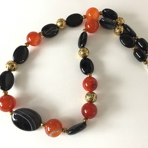 May include: A black and orange beaded necklace with gold accents. The necklace is made of oval-shaped beads and has a silver clasp.