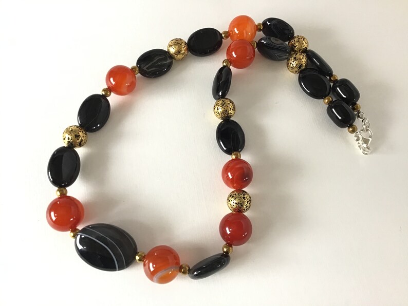 May include: A black and orange beaded necklace with gold accents. The necklace is made of oval-shaped beads and has a silver clasp.