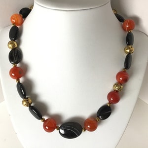 May include: A necklace made with black and orange agate beads and gold spacers. The necklace is on a white mannequin.