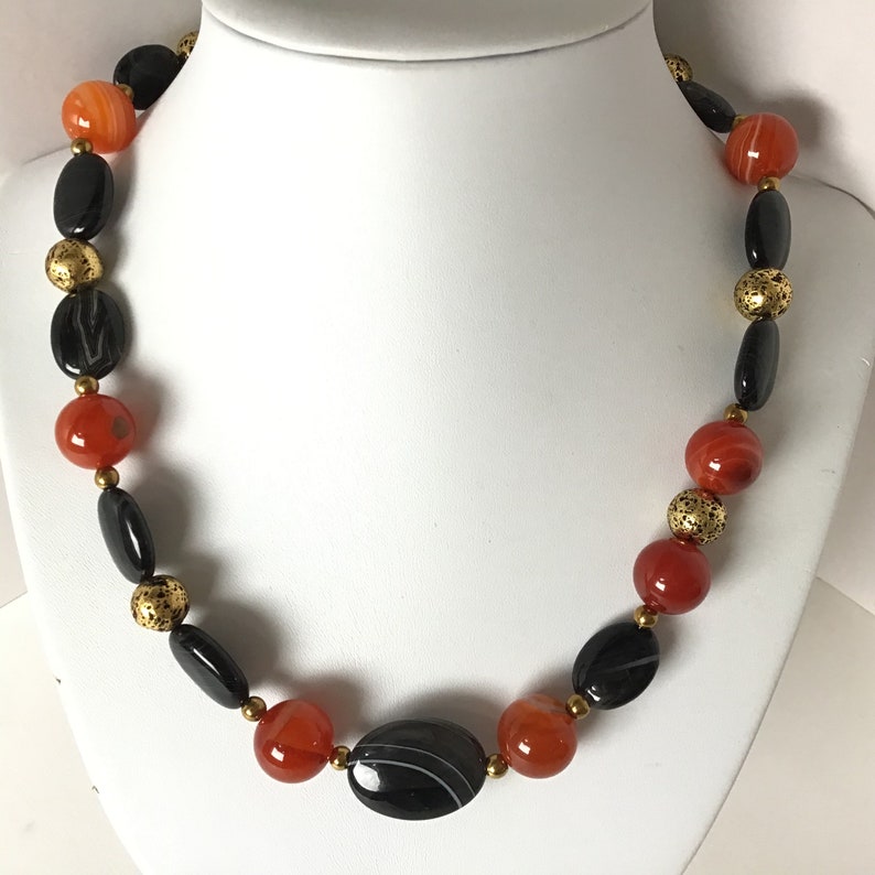 May include: A necklace made with black and orange agate beads and gold spacers. The necklace is on a white mannequin.