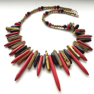 May include: A statement necklace featuring a mix of red, black, and gold beads. The necklace is made with a variety of materials, including natural stones and metal.