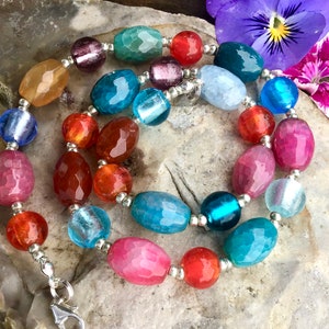 Puede incluir: Un collar de cuentas colorido con cuentas facetadas en tonos rosa, naranja, azul, verde y morado. El collar tiene un cierre plateado y se exhibe sobre una superficie de piedra natural. Las cuentas están ensartadas con metal plateado. Es una joya.