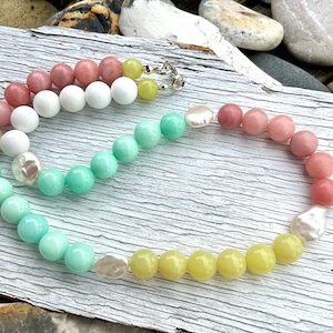 May include: A colorful beaded necklace featuring a variety of round beads in shades of pink, white, green, and yellow, interspersed with irregular white pearls. The necklace is secured with a silver clasp.