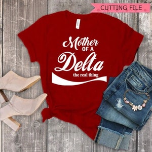 Puede incluir: Una camiseta roja con el texto blanco "Mother of a Delta the real thing". La imagen incluye vaqueros azules doblados, un collar floral y botines beige con tacón. Una pancarta rosa dice "CUTTING FILE".