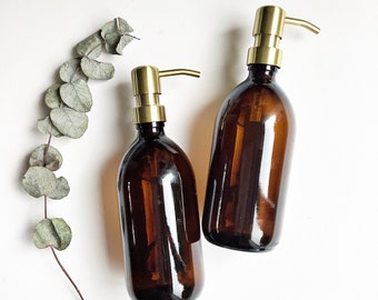 May include: Two amber glass bottles with gold pump tops. The bottles are filled with a clear liquid and are sitting on a white surface.