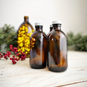 May include: Three amber glass bottles with white lids. The bottles are filled with a clear liquid. The bottles are sitting on a white wooden surface.