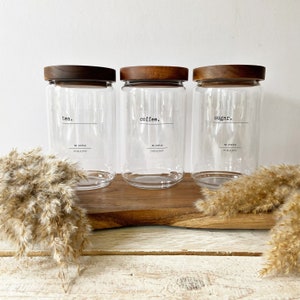 May include: Three glass canisters with wooden lids, labelled "tea", "coffee", and "sugar". The canisters are sitting on a wooden chopping board.