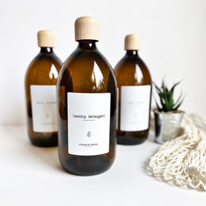 May include: Three amber glass bottles with wooden lids and white labels. The centre bottle label reads "laundry detergent" with a small floral design and the text "CHARLIE ROSE" below.