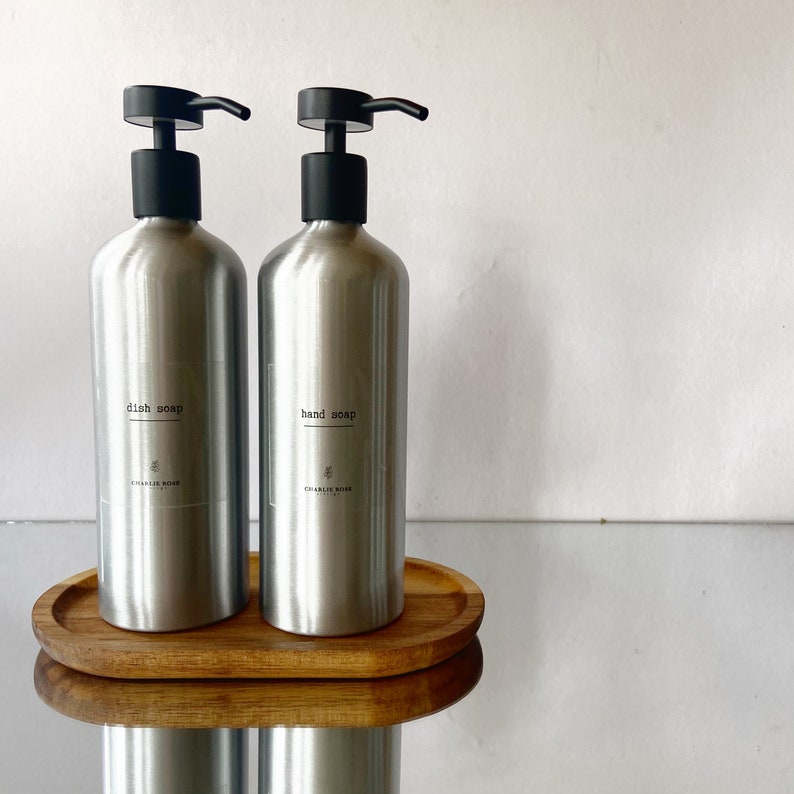 May include: Two silver pump bottles with black pumps, one labelled "dish soap" and the other "hand soap", both with the text "CHARLIE ROSE" on the label, sitting on a wooden tray.