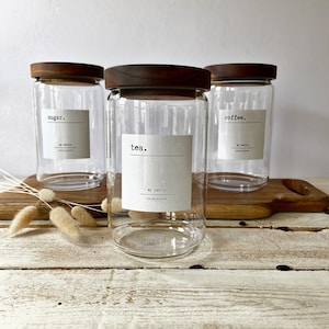 May include: Three glass jars with wooden lids and labels. The labels read "sugar", "tea", and "coffee". The jars are sitting on a wooden surface.