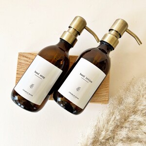 May include: Two amber glass bottles with gold pumps and white labels. The labels read "hand soap" and "hand lotion" with the text "CHARLIE ROSE" underneath. The bottles are sitting on a wooden surface.