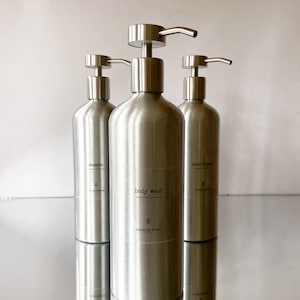 May include: Three silver pump bottles with labels reading "shampoo", "body wash", and "conditioner". The bottles are on a reflective surface.