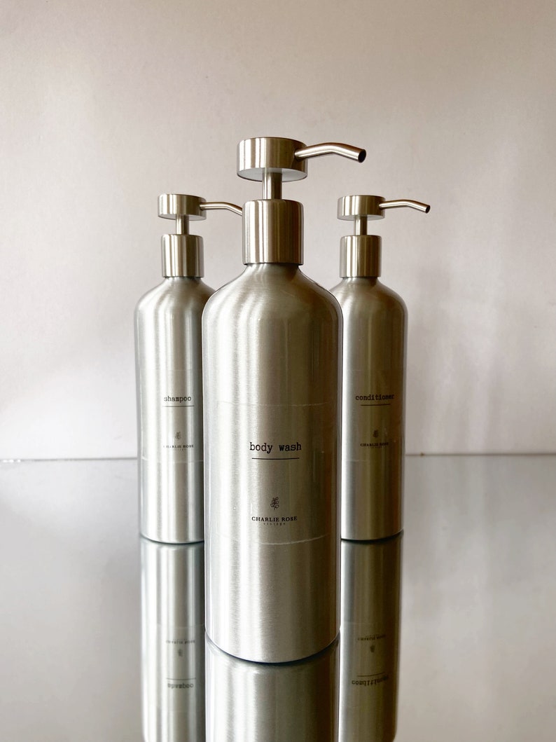 May include: Three silver pump bottles with labels reading "shampoo", "body wash", and "conditioner". The bottles are on a reflective surface.