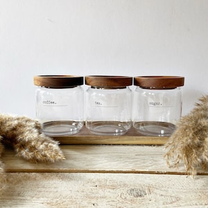 May include: Three glass jars with wooden lids on a wooden tray. The jars are labelled "coffee", "tea", and "sugar".