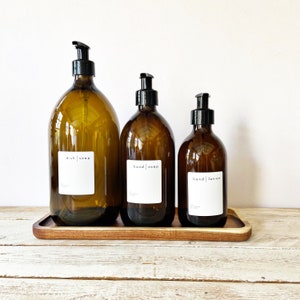 May include: Three amber glass bottles with black pump tops and white labels. The labels read "dish soap", "hand soap", and "hand lotion". The bottles are sitting on a wooden tray.